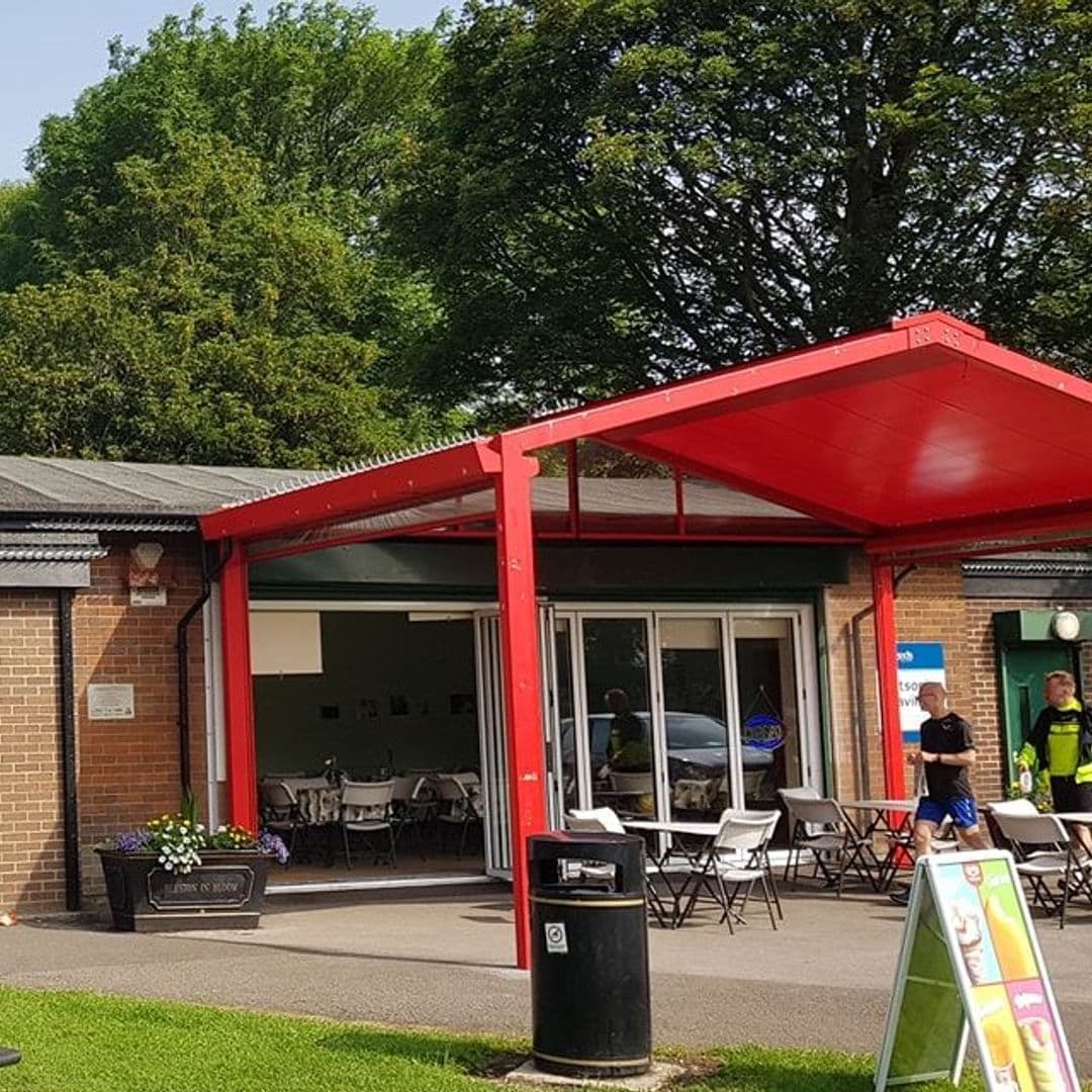 Cross_Flatts_Park_Cafe_Sunny_Day_Outdoor_Seating Exterior view of Cross Flatts Park Cafe with red awning, outdoor seating, and lush green trees in the background.