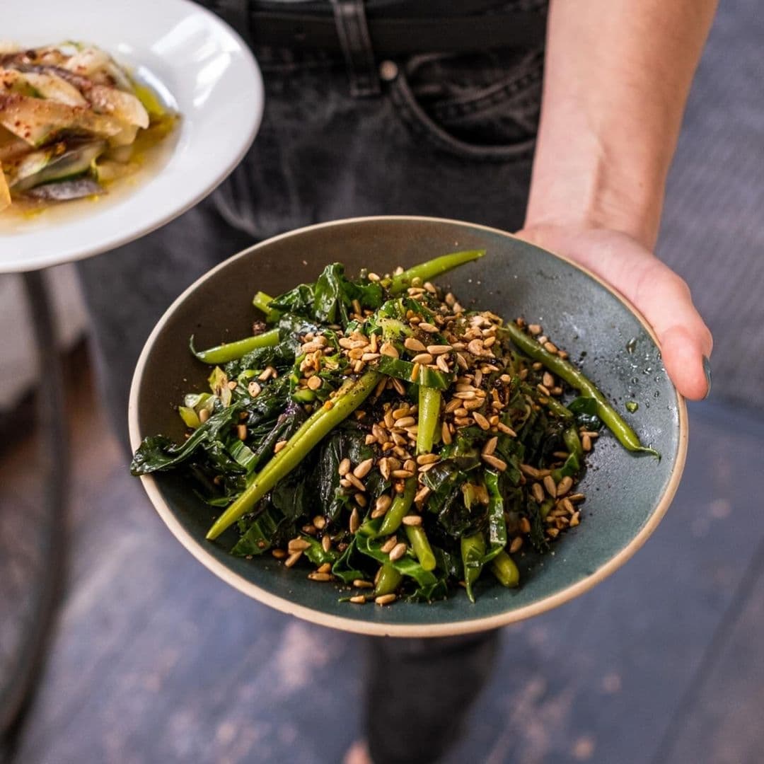 Eat Your Greens Fresh Sautéed Vegetables Seed Garnish A person holding a bowl of fresh green vegetables topped with seeds at Eat Your Greens.