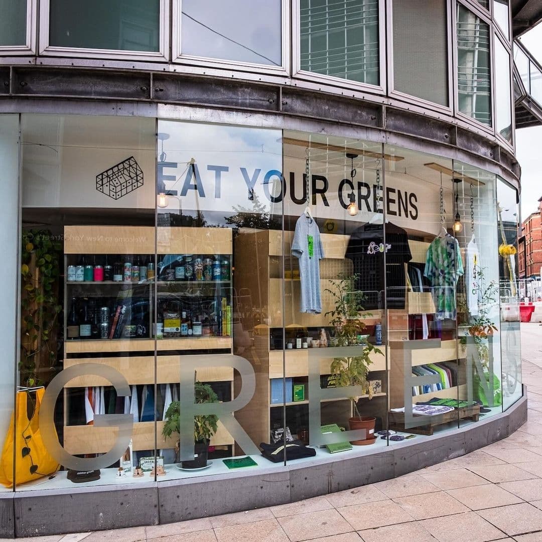 Eat Your Greens Storefront Eco Friendly Products Display Corner storefront of "Eat Your Greens" with large windows displaying various plants, books, clothing, and eco-friendly products.