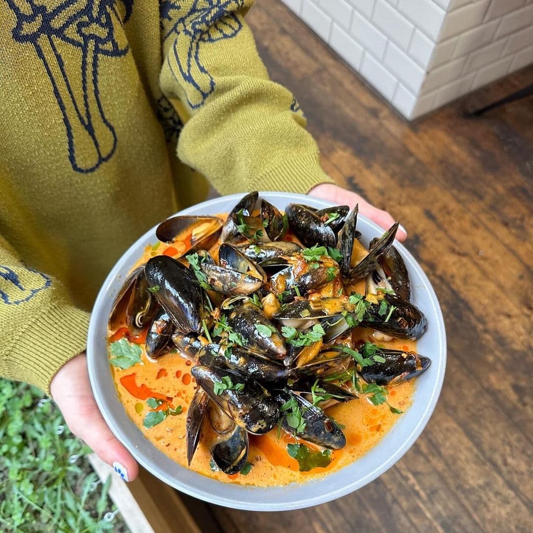 EatYourGreens_FreshMussels_DeliciousSeafood_CulinaryExperience Person holding a bowl of mussels garnished with herbs at Eat Your Greens.
