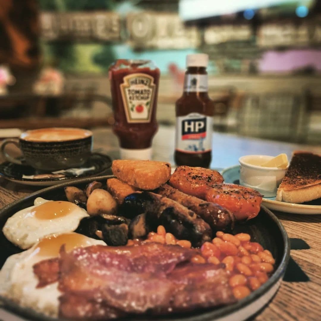 Editors Draught Full English Breakfast Coffee Condiments Traditional English breakfast served at Editor's Draught with eggs, bacon, sausages, beans, mushrooms, hash browns, and toast, accompanied by a cup of coffee and condiments.