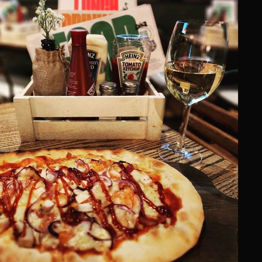 Editors Draught Gourmet Pizza Wine Dining Experience Alt text: "Enjoy a delicious pizza with toppings and drizzled sauce paired with a glass of white wine at Editor’s Draught. Condiments and a small flower decoration in the background add to the cozy dining atmosphere."