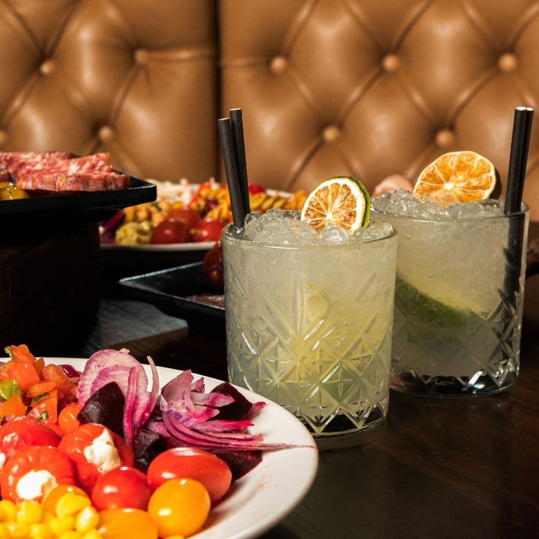 Estabulo The Light Cocktails Fine Dining Leather Booths Two refreshing cocktails with lime garnish served at Estabulo – The Light, accompanied by a colorful plate of salad and a selection of meats in the background, presented on a wooden table with a tufted leather booth backdrop.