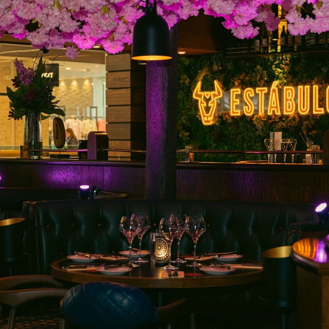 Estabulo The Light Elegant Dining Ambiance Floral Decor Interior of Estabulo – The Light featuring an intimate dining setup with wine glasses on the table, ambient lighting, and vibrant pink floral decor overhead. A neon sign with the restaurant's name is visible in the background.