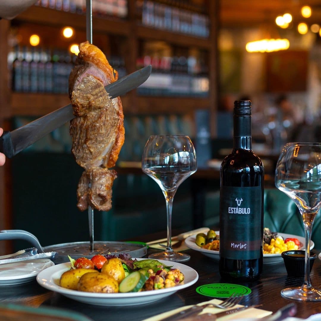 Estabulo The Light Grilled Meat Wine Dining Experience Grilled meat on a skewer being served at Estabulo – The Light, with a bottle of Estabulo Merlot wine and empty wine glasses on a table set with various dishes in a restaurant setting.