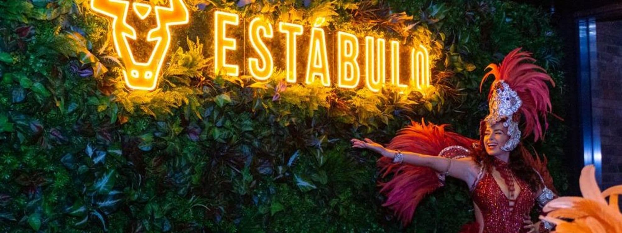 Performer in vibrant carnival costume at Estabulo – The Light with neon sign in background.