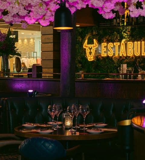 Estabulo The Light_Elegant Dining Ambiance Floral Decor Elegant dining setup at Estabulo – The Light, featuring a cozy booth with wine glasses and plates, under ambient lighting with vibrant pink flowers overhead and the restaurant's glowing sign in the background.