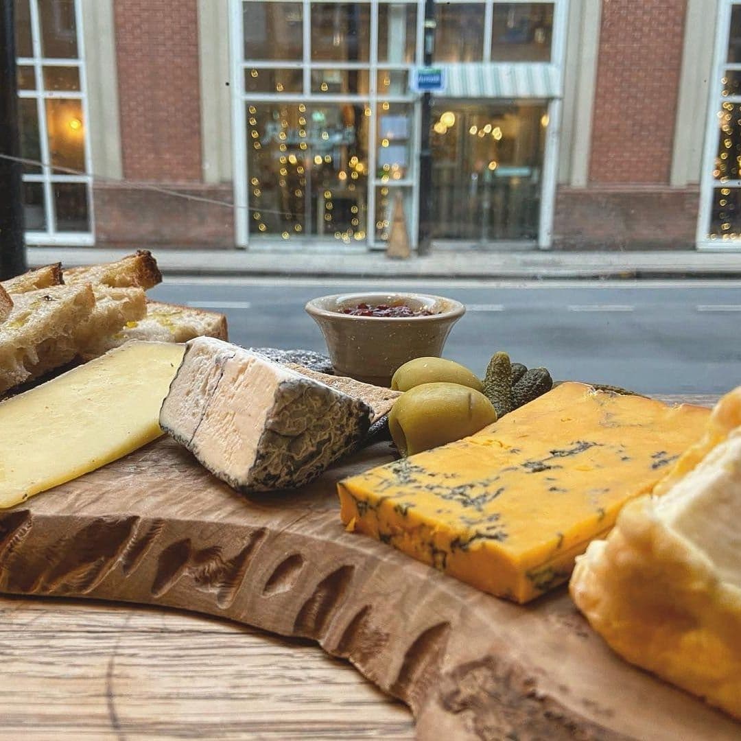 Friends of Ham Leeds City Artisan Cheese Platter Window View "Assorted cheeses and bread on a wooden board at Friends of Ham – Leeds City, with a street view through a window in the background."