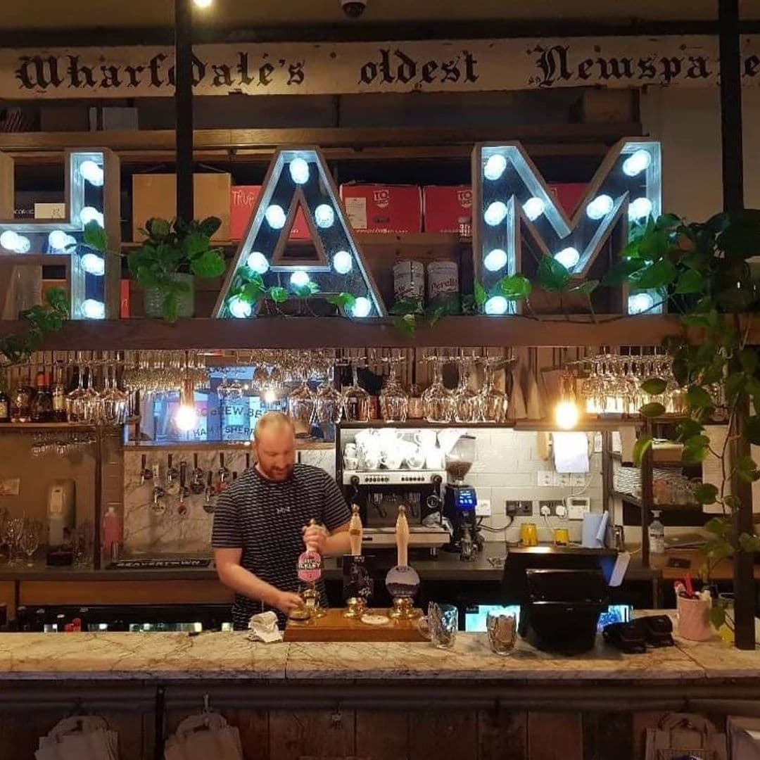 Friends of Ham Leeds City Bartender Pouring Drink Neon Signs Barman pouring a drink at Friends of Ham – Leeds City, with illuminated 'HAM' letters on the shelf and a variety of glasses hanging above the bar.