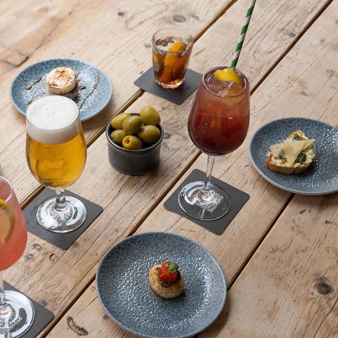 Friends of Ham Leeds City Gourmet Tapas Craft Beer Cocktails Assorted gourmet tapas and drinks on a wooden table at Friends of Ham – Leeds City, featuring a glass of beer, a cocktail with a straw, a small bowl of olives, and various small plates with appetizers.