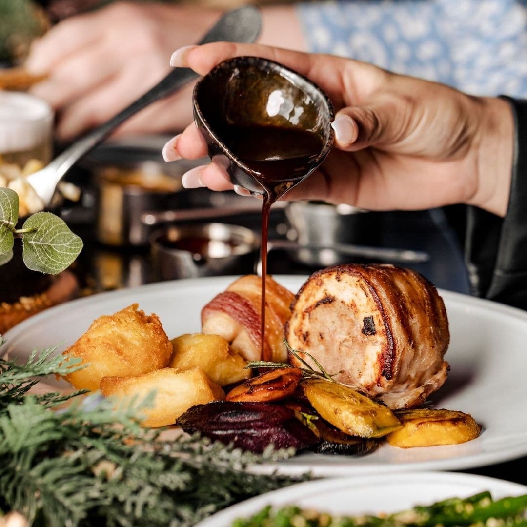 Gaucho Traditional Cuisine Pouring Sauce Roasted Meat Alt text: "Savor the flavors of Gaucho with a dish featuring succulent roasted meat wrapped in bacon, served with golden potatoes, roasted vegetables, and a rich, dark sauce being poured over."