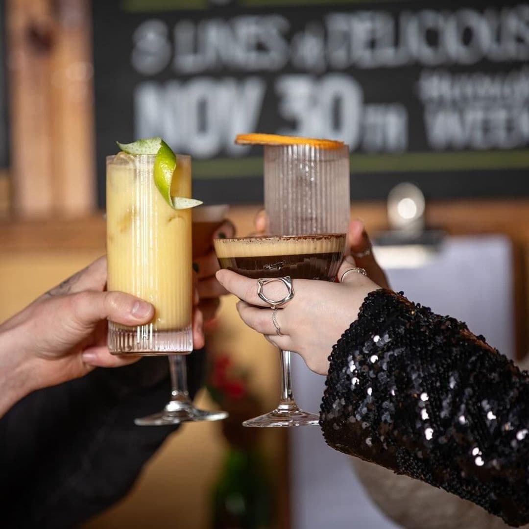 Granvilles Beer Gin House Cocktail Cheers Celebration Two hands toasting with cocktails at Granvilles Beer & Gin House, one holding a tall glass with a yellow drink and lime wedge, the other a stemmed glass with a layered coffee-flavored cocktail, garnished with an orange slice.