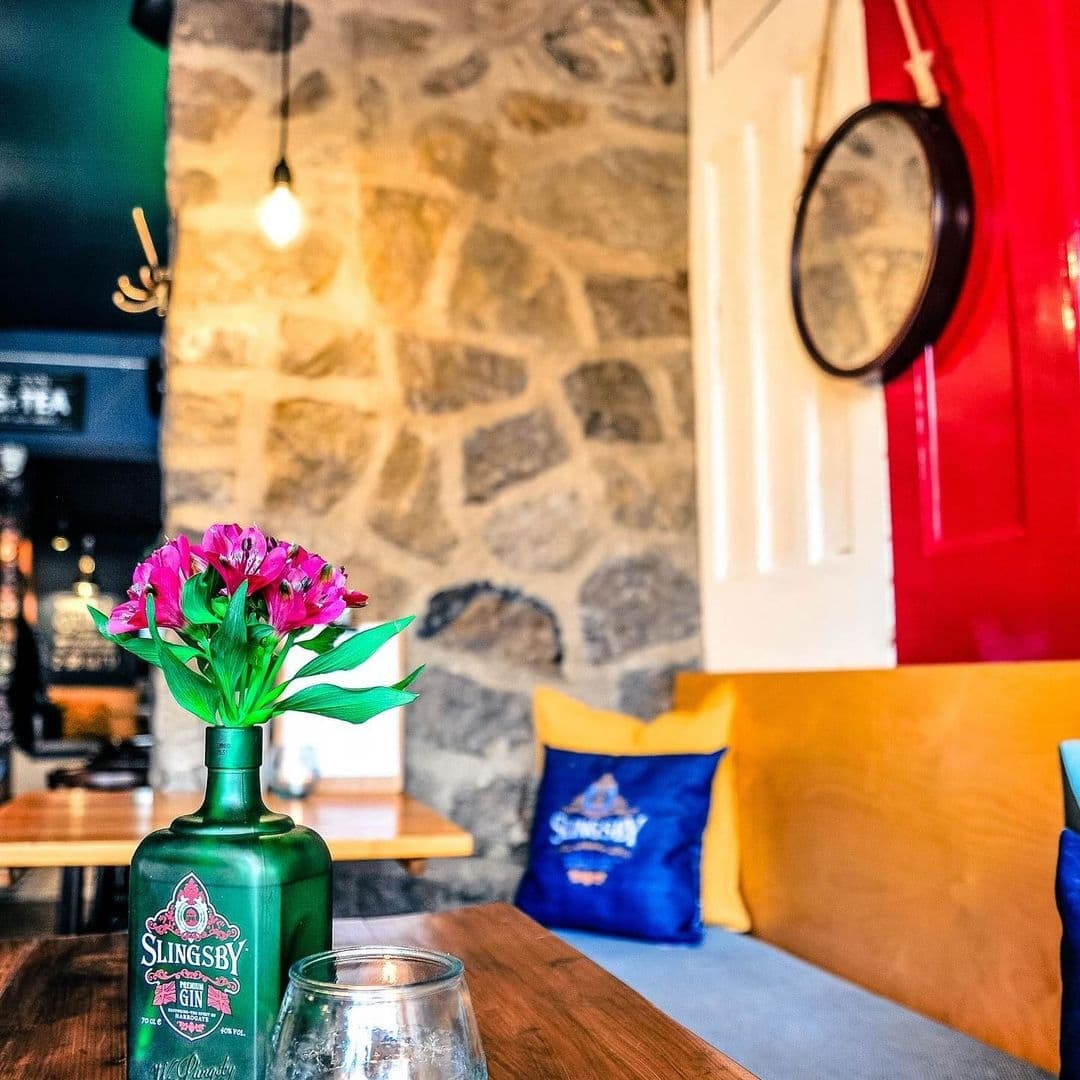 Granvilles Beer Gin House Cozy Interior Flowers Decor Cozy corner at Granvilles Beer & Gin House with a vibrant bouquet of pink flowers in a green Slingsby Gin bottle, stone walls, and comfortable seating with yellow cushions.