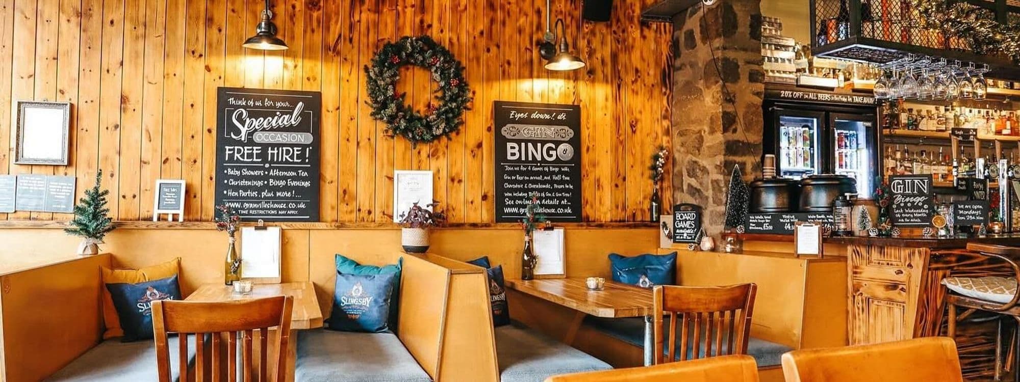 Interior of Granvilles Beer & Gin House featuring wooden walls with festive decorations, cozy seating areas with cushions, a bar with various drinks on display, and signage advertising events and specials.