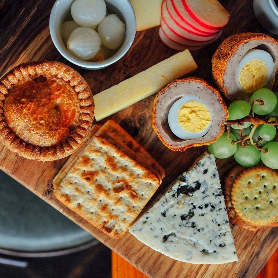 Granvilles Beer Gin House Gourmet Platter Cheese Scotch Egg Alt text: "A selection of savory snacks at Granvilles Beer & Gin House, featuring a golden-brown meat pie, hard cheeses, a scotch egg, crackers, pickled onions, radish slices, and green grapes, all arranged on a wooden serving board."