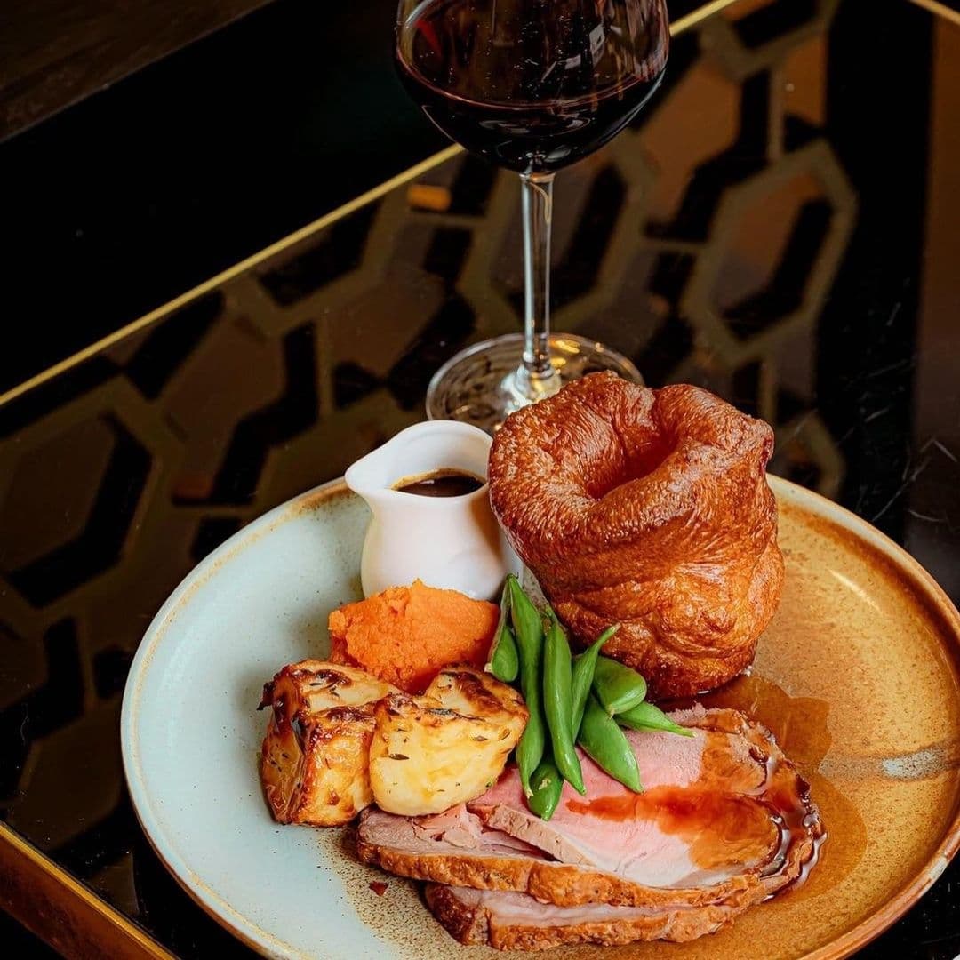 HARVEY NICHOLS Fourth Floor Brasserie Bar Gourmet Roast Dinner Wine Gourmet roast dinner with succulent slices of meat, golden Yorkshire pudding, roasted potatoes, green beans, carrot puree, and gravy, accompanied by a glass of red wine at Harvey Nichols Fourth Floor Brasserie & Bar.