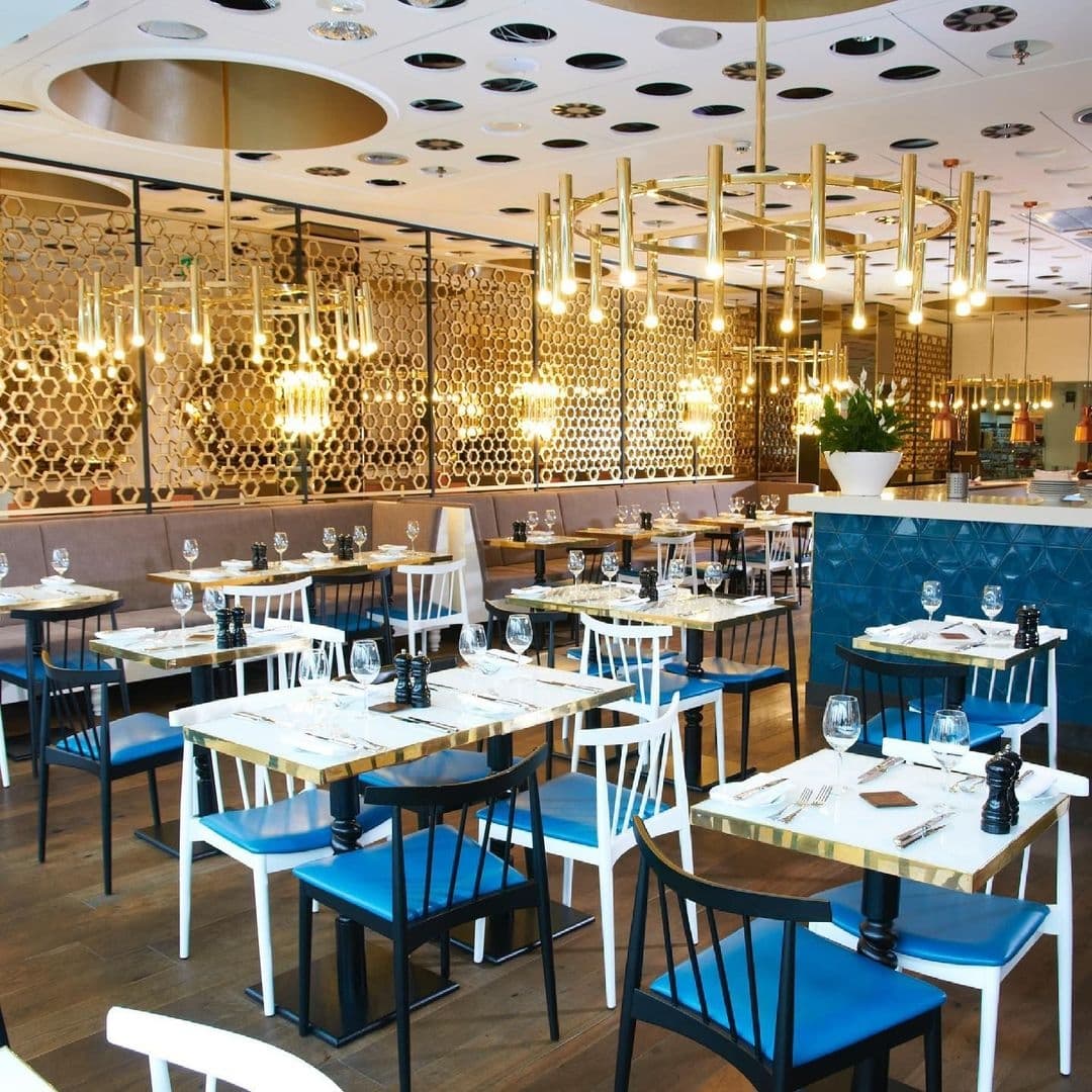 HARVEY NICHOLS Fourth Floor Brasserie Elegant Interior Design Elegant dining area at Harvey Nichols Fourth Floor Brasserie & Bar with modern decor, patterned golden wall panels, stylish hanging lights, and neatly set tables with blue and white chairs.