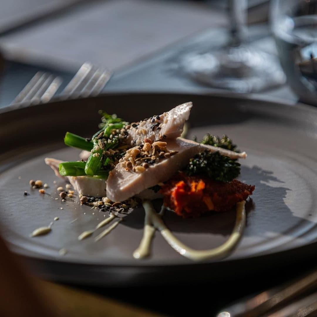 HARVEY NICHOLS Fourth Floor Brasserie Gourmet Dish Presentation Gourmet dish served at H A R V E Y • N I C H O L S • Fourth Floor • Brasserie & Bar, featuring slices of chicken on a bed of green vegetables, sprinkled with seeds and nuts, accompanied by a dollop of red sauce and a creamy drizzle on a dark plate.