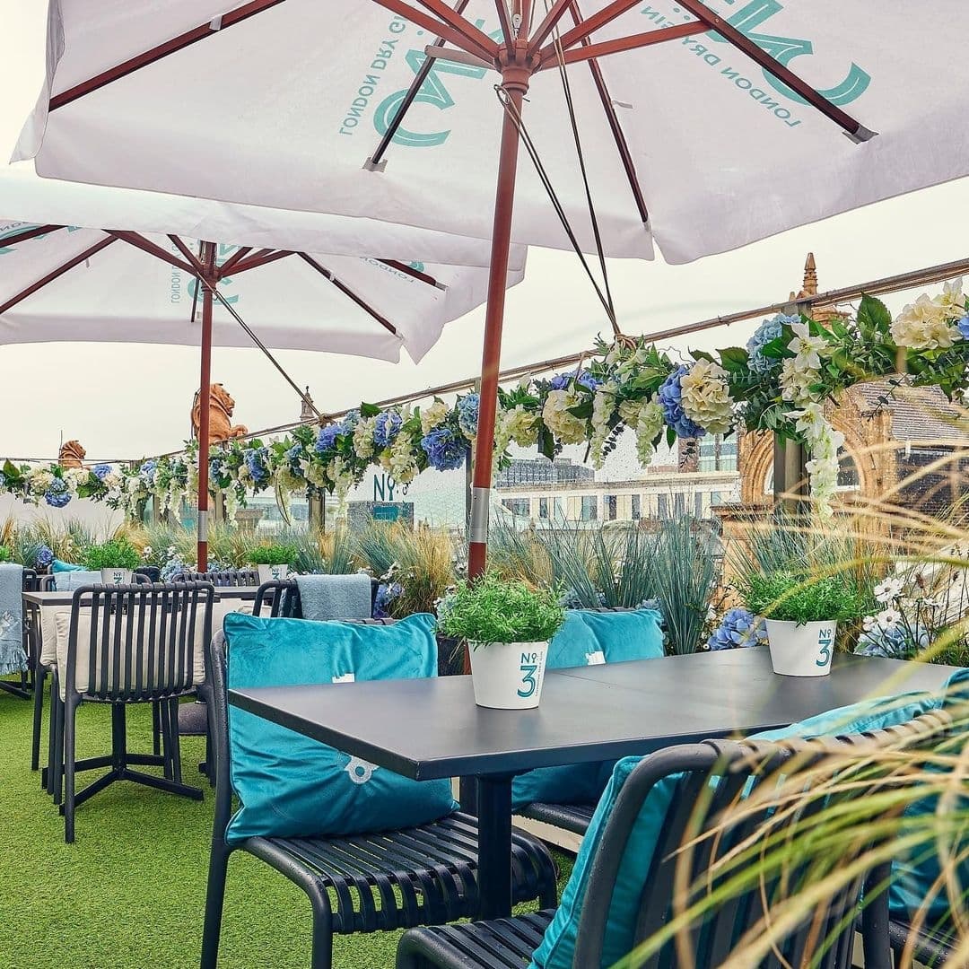 HARVEY NICHOLS Fourth Floor Brasserie Umbrellas Outdoor Seating Floral Decor Outdoor seating area at H A R V E Y • N I C H O L S • Fourth Floor • Brasserie & Bar with comfortable blue cushions, black chairs, and tables under white umbrellas, surrounded by green plants and decorative flowers.