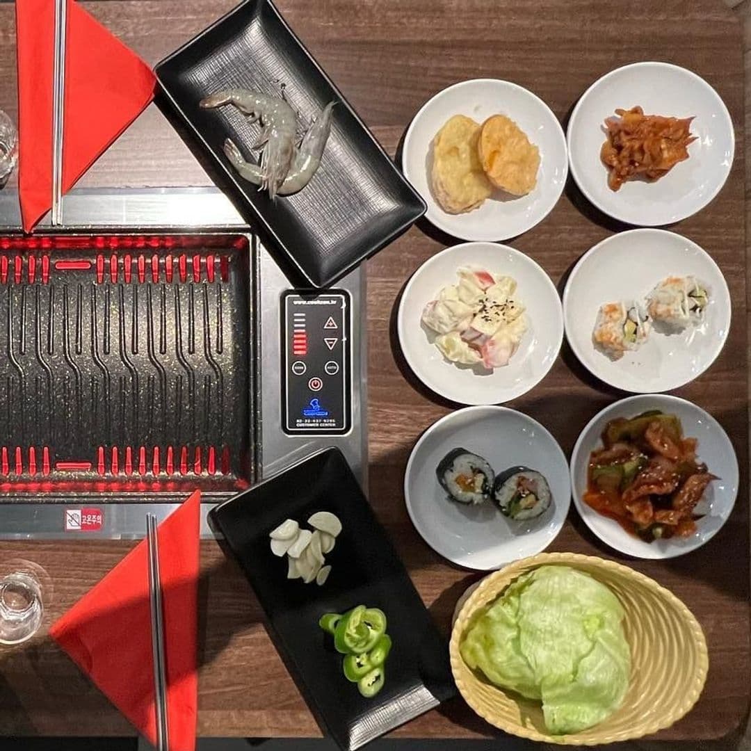 Hama Korean BBQ Buffet Assorted Dishes Grill Table Setting Tabletop with a variety of Korean dishes at Hama Korean BBQ Buffet, featuring a grill ready for cooking, plates with shrimp, sushi rolls, dumplings, kimchi, and fresh lettuce, with sliced garlic and green chili peppers on the side.