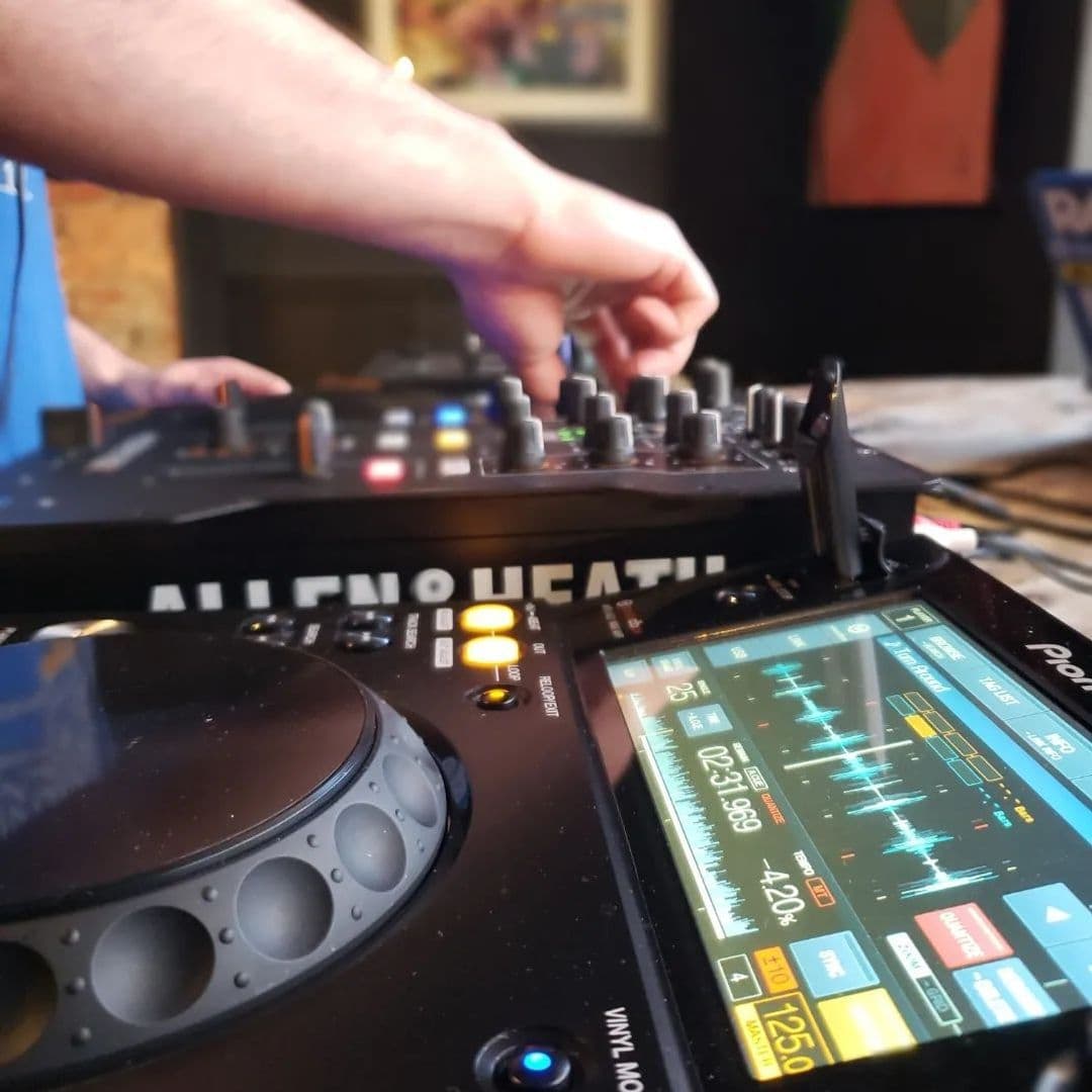 Handsome Bar DJ Mixing Deck Live Music Close-up of a DJ's hand adjusting controls on a mixing console at Handsome Bar, with a blurred background suggesting a lively atmosphere.