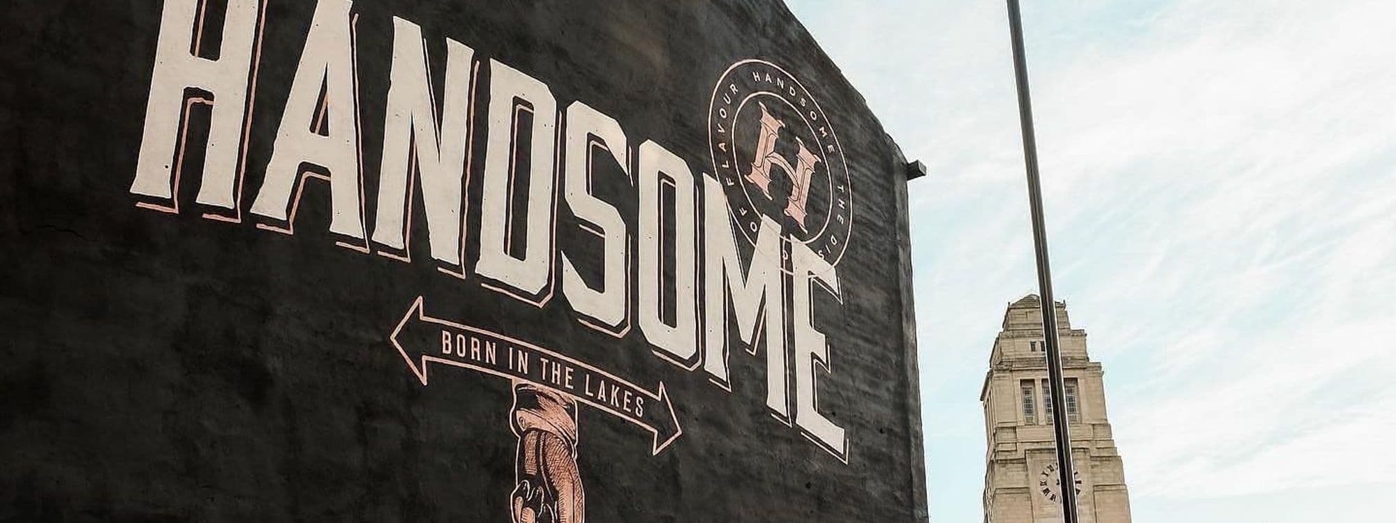 Exterior wall of Handsome Bar with large mural-style signage, featuring bold white letters on a black background and an illustration of a hand holding a drink, with the tagline "Born in the Lakes" beneath.