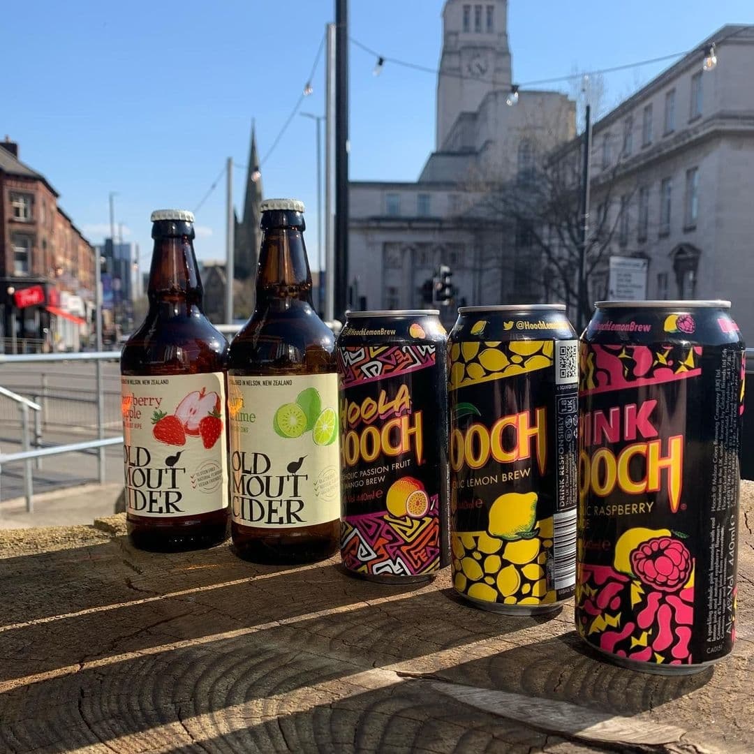 Handsome_Bar_Craft_Beers_Sunny_Day_Outdoor Assorted bottles and cans of cider and kombucha on a wooden table at Handsome Bar with a city street and buildings in the background.