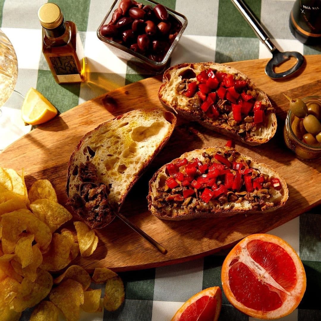 Harvey Nichols Fourth Floor Brasserie Delicious Bruschetta Platter Gourmet open-faced sandwiches with vibrant red bell pepper topping, served on a wooden board with crisps, olives, and a side of fresh grapefruit, at H A R V E Y • N I C H O L S • Fourth Floor • Brasserie & Bar.