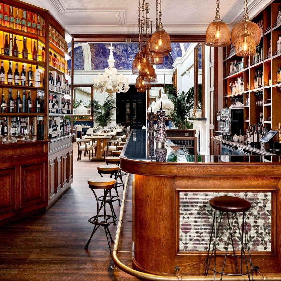 Ibérica Elegant Bar Interior Wooden Furnishings Vintage Lighting Elegant interior of Ibérica featuring a curved wooden bar with pendant lights, bar stools, and shelves stocked with wine bottles, leading to a dining area with tables set for service.
