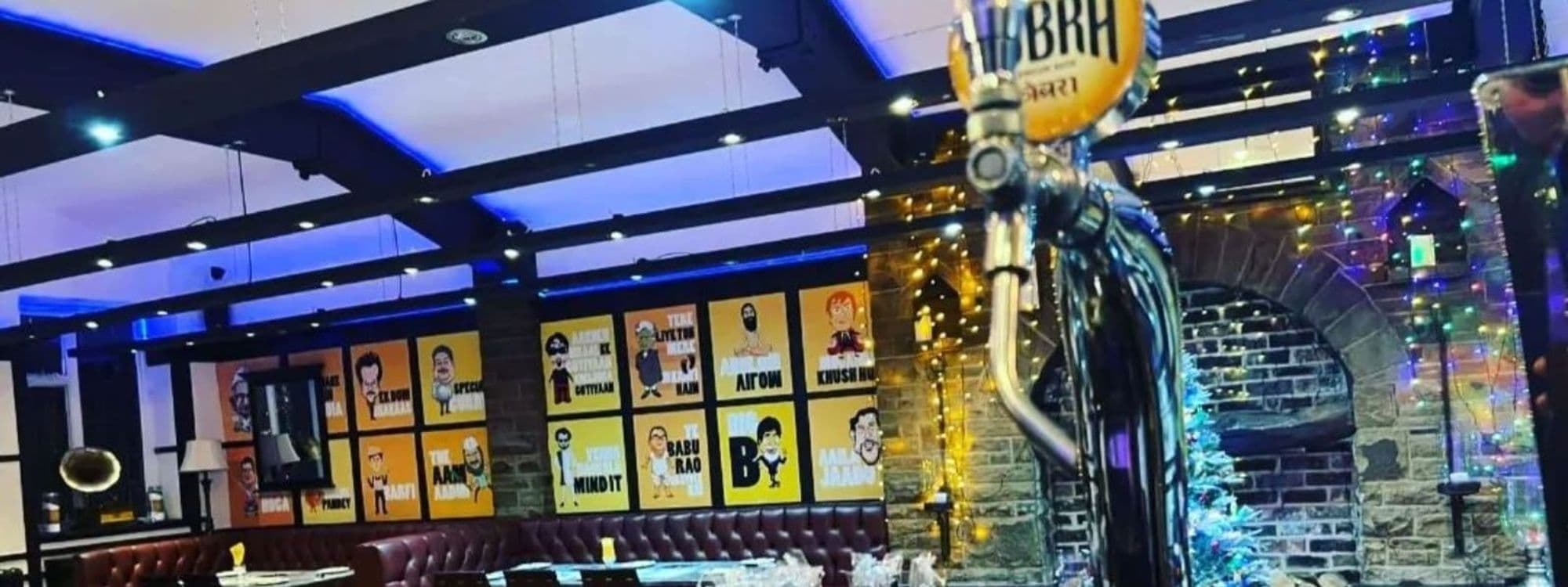 Interior of Indian Pakwaan restaurant showing a bar with a beer tap in the foreground, colorful pop art style portraits on the wall, cozy booth seating, and festive string lights.