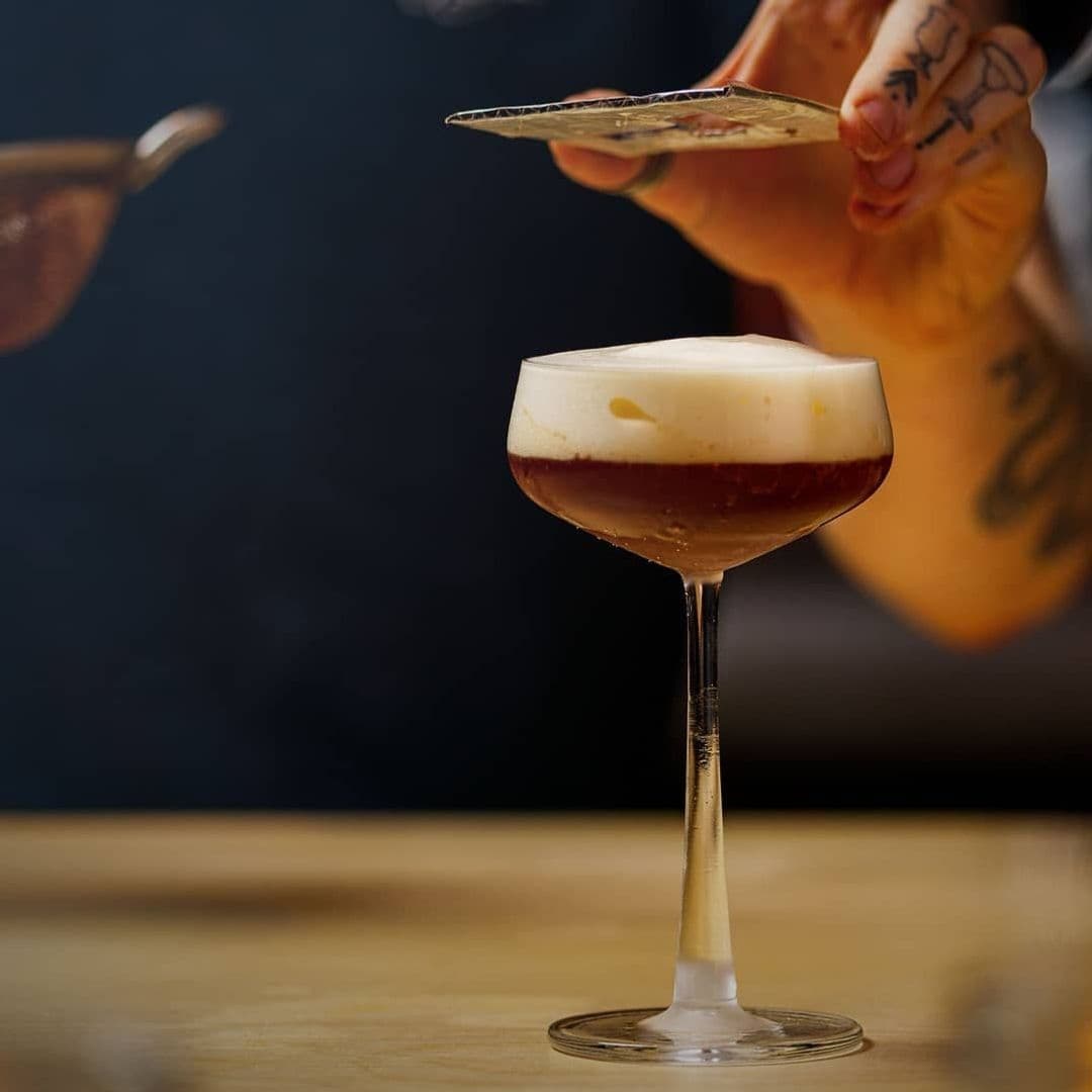 Jakes Bar Still Room Craft Cocktail Pouring Experience Craft cocktail with a frothy top served at Jake's Bar & Still Room, with a bartender's tattooed hand in the background.