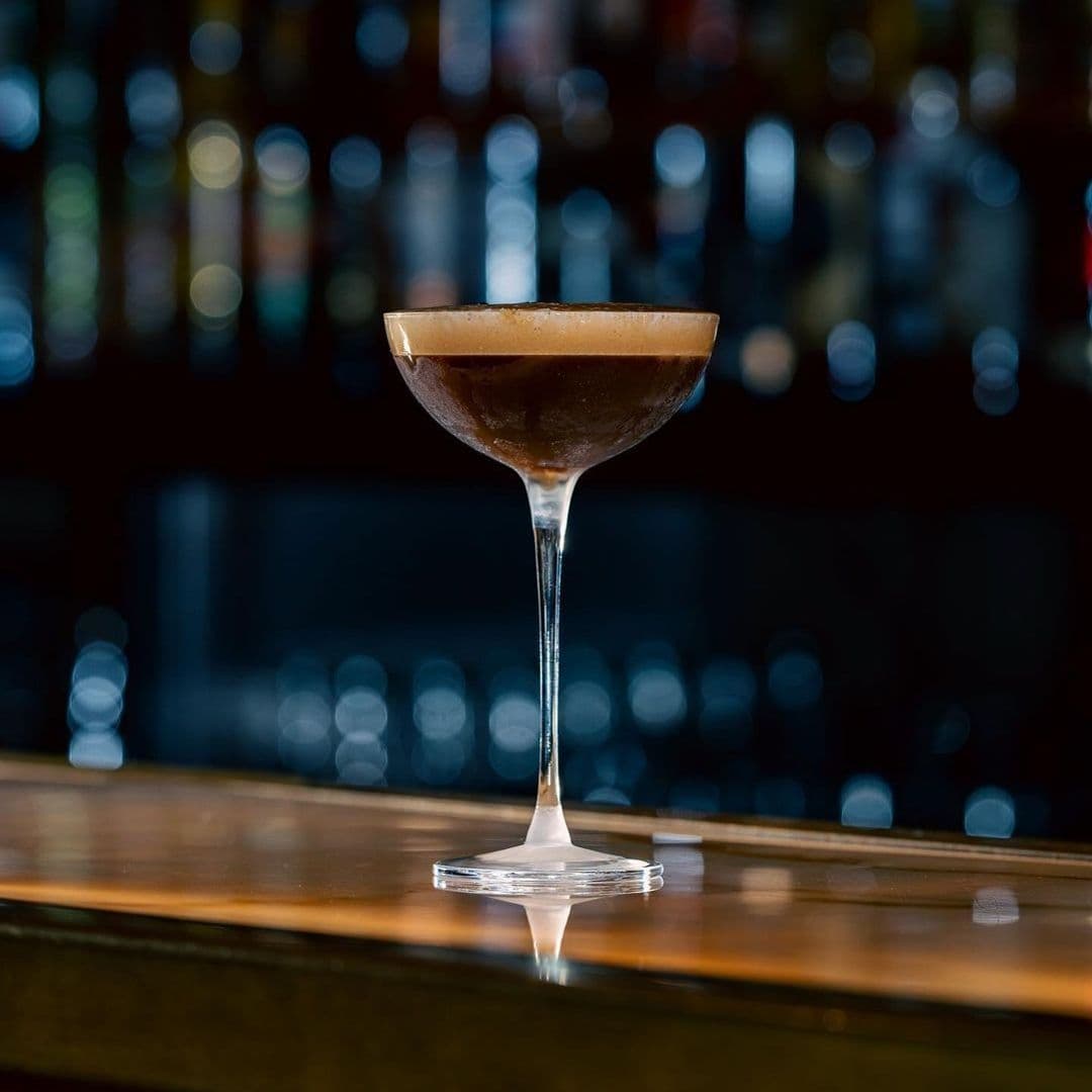 Jakes Bar Still Room Elegant Cocktail Glass Bokeh Background Espresso martini served at Jake's Bar & Still Room, with a frothy top, in a classic cocktail glass on a wooden bar counter, backlit by a blurred array of bottles.