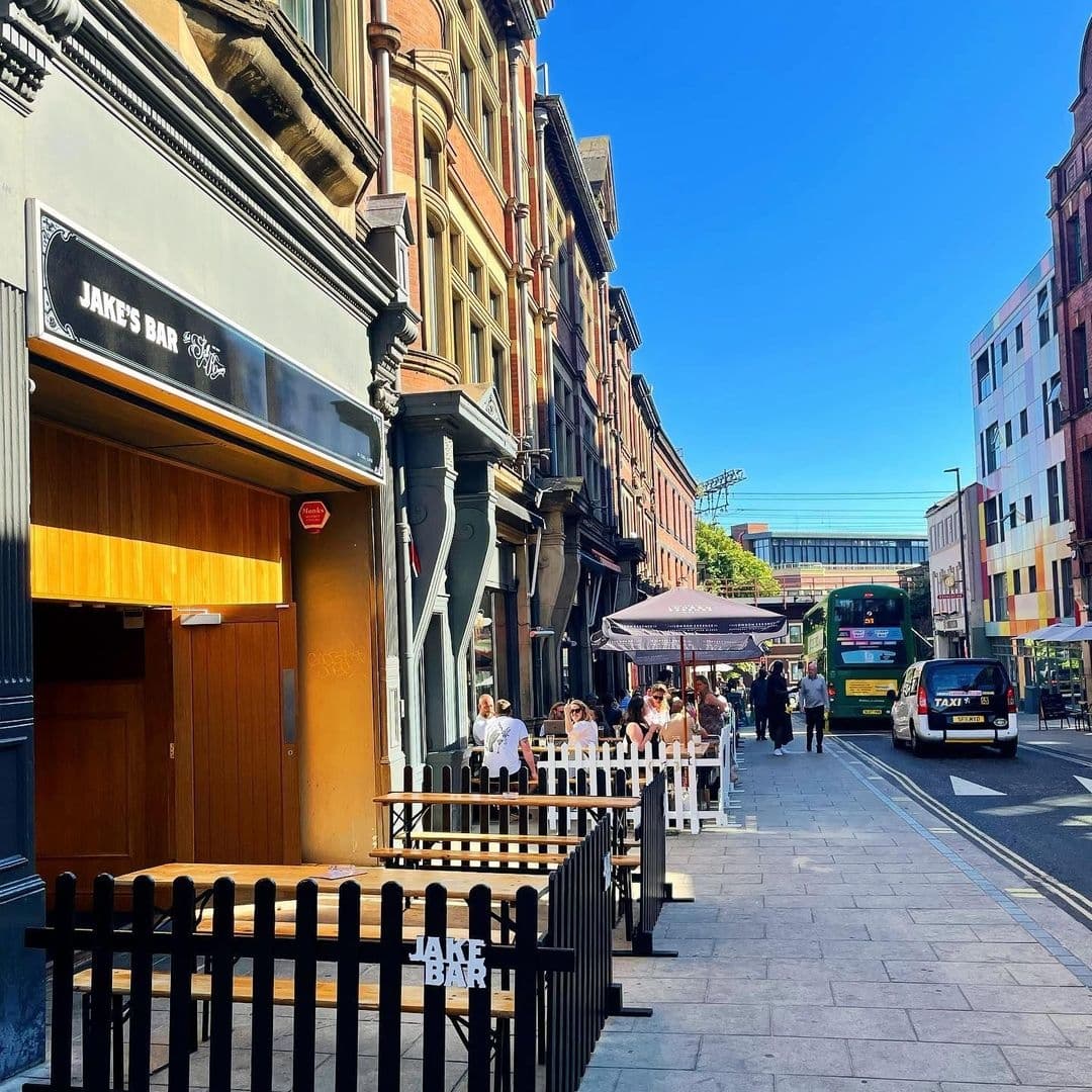 Jakes Bar Still Room Sunny Street Scene Social Gathering Exterior view of Jake's Bar & Still Room with patrons seated outside on a sunny day, city street scene with a bus and taxi passing by.