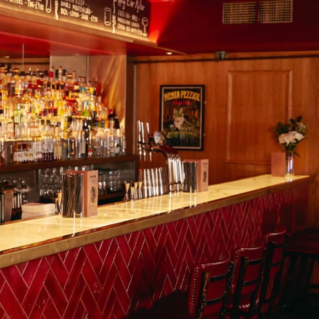 Jakes Bar Still Room cozy interior bar counter ambient lighting Interior of Jake's Bar & Still Room featuring a polished bar counter with red diamond-patterned front, wooden stools, and a well-stocked bar with a variety of bottles. A vintage poster adds character to the warm, inviting ambiance.