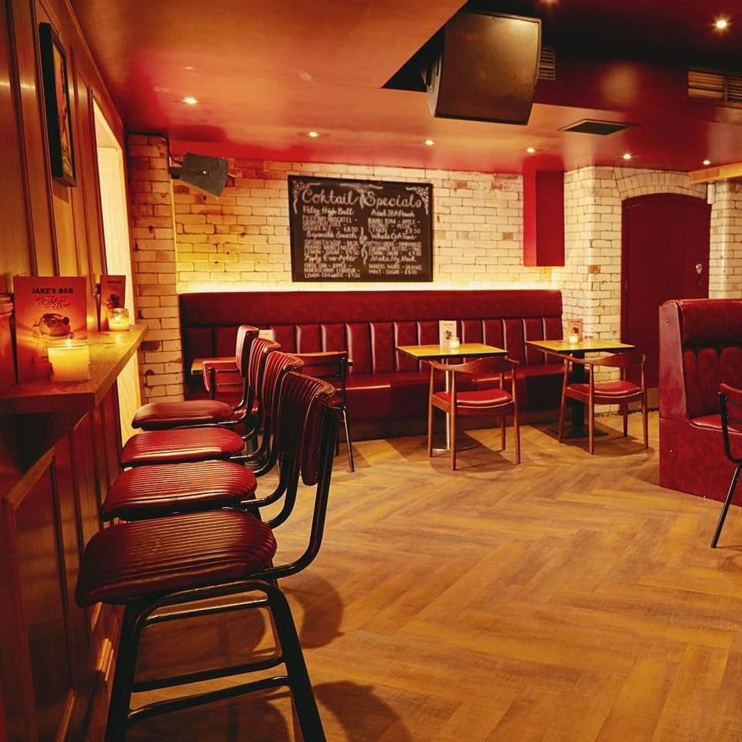 Jakes Bar Still Room cozy interior brick walls wooden floor Interior of Jake's Bar & Still Room featuring a cozy seating area with red upholstered booths, wooden tables, and bar stools lined up along a wooden counter. Warm lighting and exposed brick walls create an inviting atmosphere, with a chalkboard displaying cocktail specials in the background.