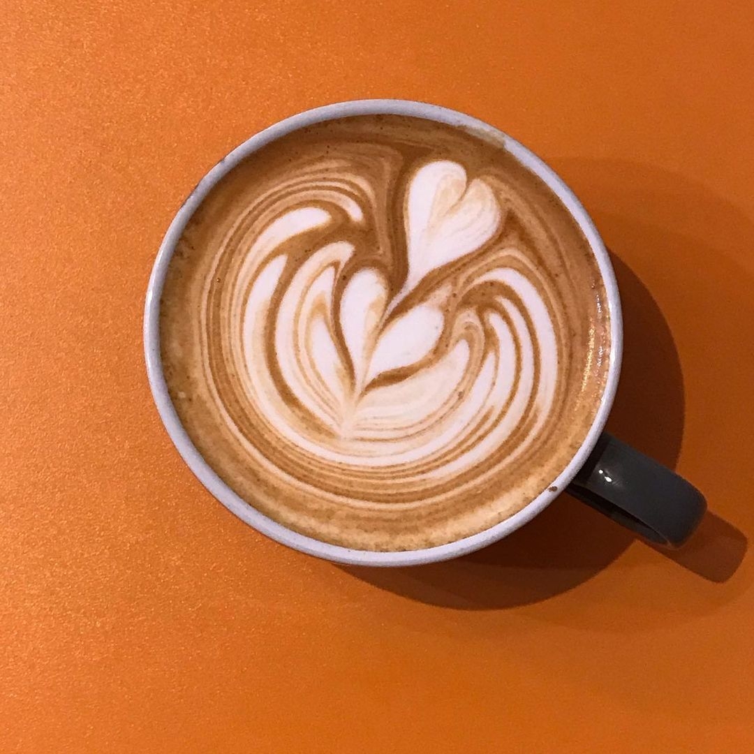 Kapow Coffee The Calls Artistic Latte Orange Background Artistic latte with a heart-shaped design at Kapow Coffee – The Calls, on an orange background.