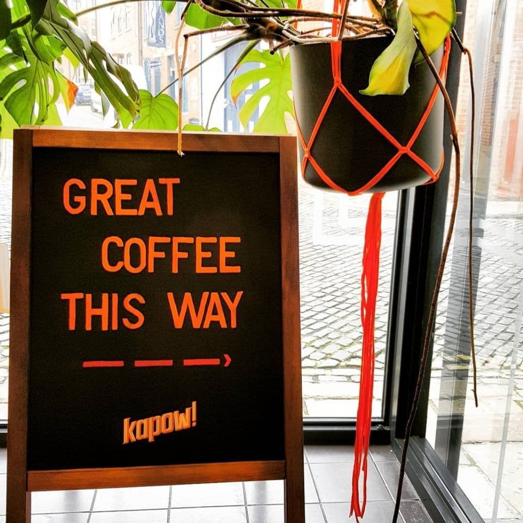Kapow Coffee The Calls Great Coffee Sign Inviting Entrance "Inviting signboard at Kapow Coffee – The Calls indicating 'GREAT COFFEE THIS WAY' with an arrow, set against a window view with a hanging plant and cobblestone street outside."