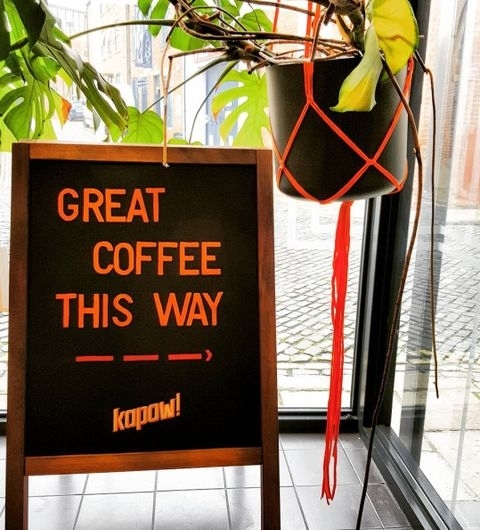 Kapow Coffee The Calls Great Coffee Sign Window Plant Alt text: "Signboard at Kapow Coffee – The Calls indicating 'GREAT COFFEE THIS WAY' with an arrow and the Kapow logo, next to a potted plant by a window."