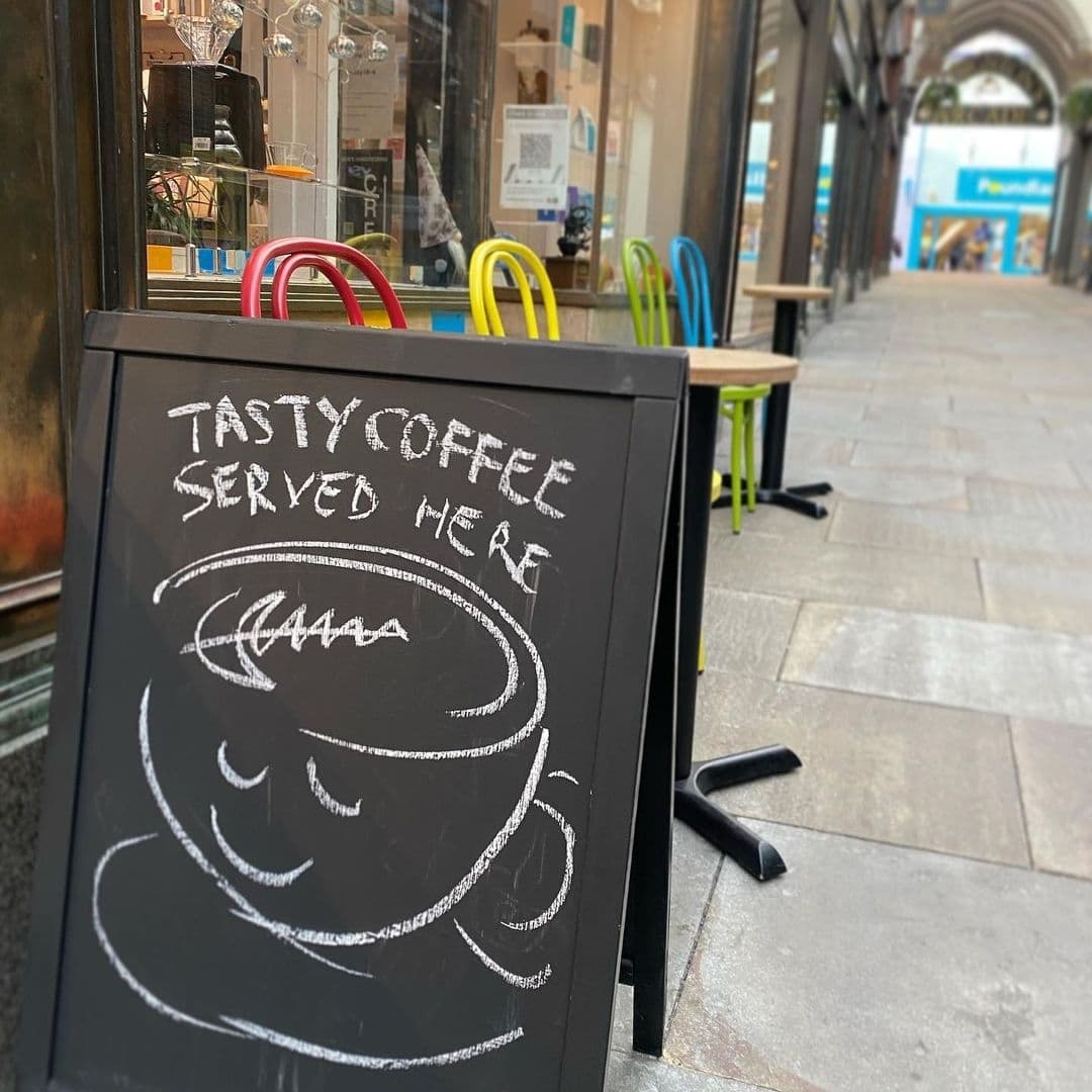 Kapow Coffee Thorntons Arcade Coffee Sign Arcade View A sidewalk sign with the words "TASTY COFFEE SERVED HERE" and a drawing of a steaming coffee cup in front of Kapow Coffee – Thorntons Arcade, with colorful chairs in the background.