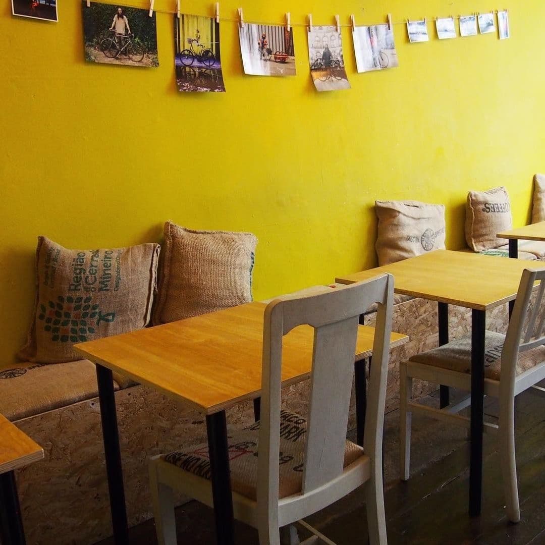 Kapow Coffee Thorntons Arcade cozy interior yellow wall photo display Interior of Kapow Coffee – Thorntons Arcade with wooden tables, white chairs, and burlap cushioned benches against a vibrant yellow wall adorned with hanging photographs.