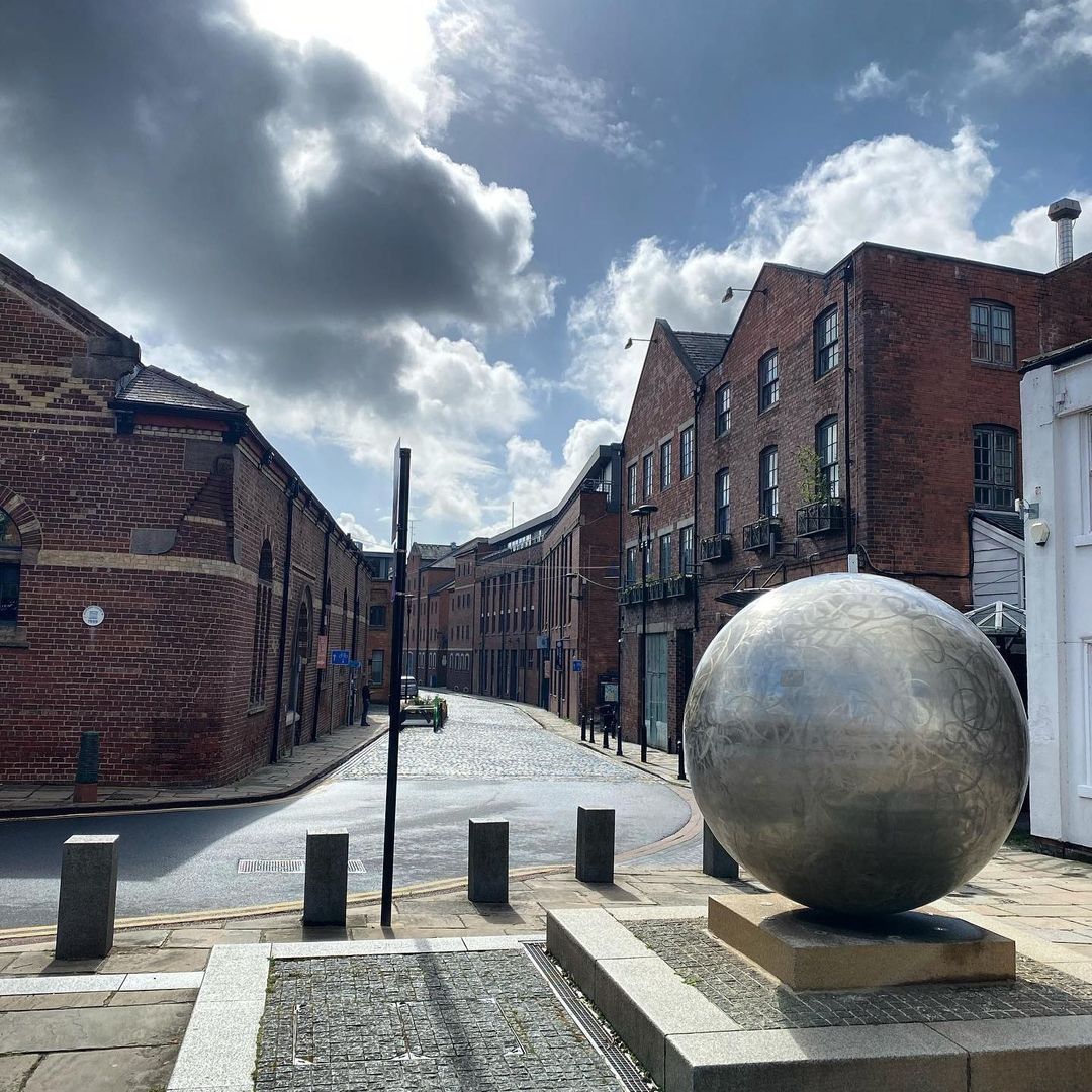 Kapow_Coffee_The_Calls_Urban_Street_Sphere_Sculpture Cobbled street leading to Kapow Coffee – The Calls, featuring a large metallic sphere sculpture, with historic brick buildings under a partly cloudy sky.
