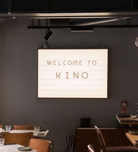 Kino_Welcome_Sign_Restaurant_Interior Interior of Kino with a welcome sign above wooden tables and chairs.