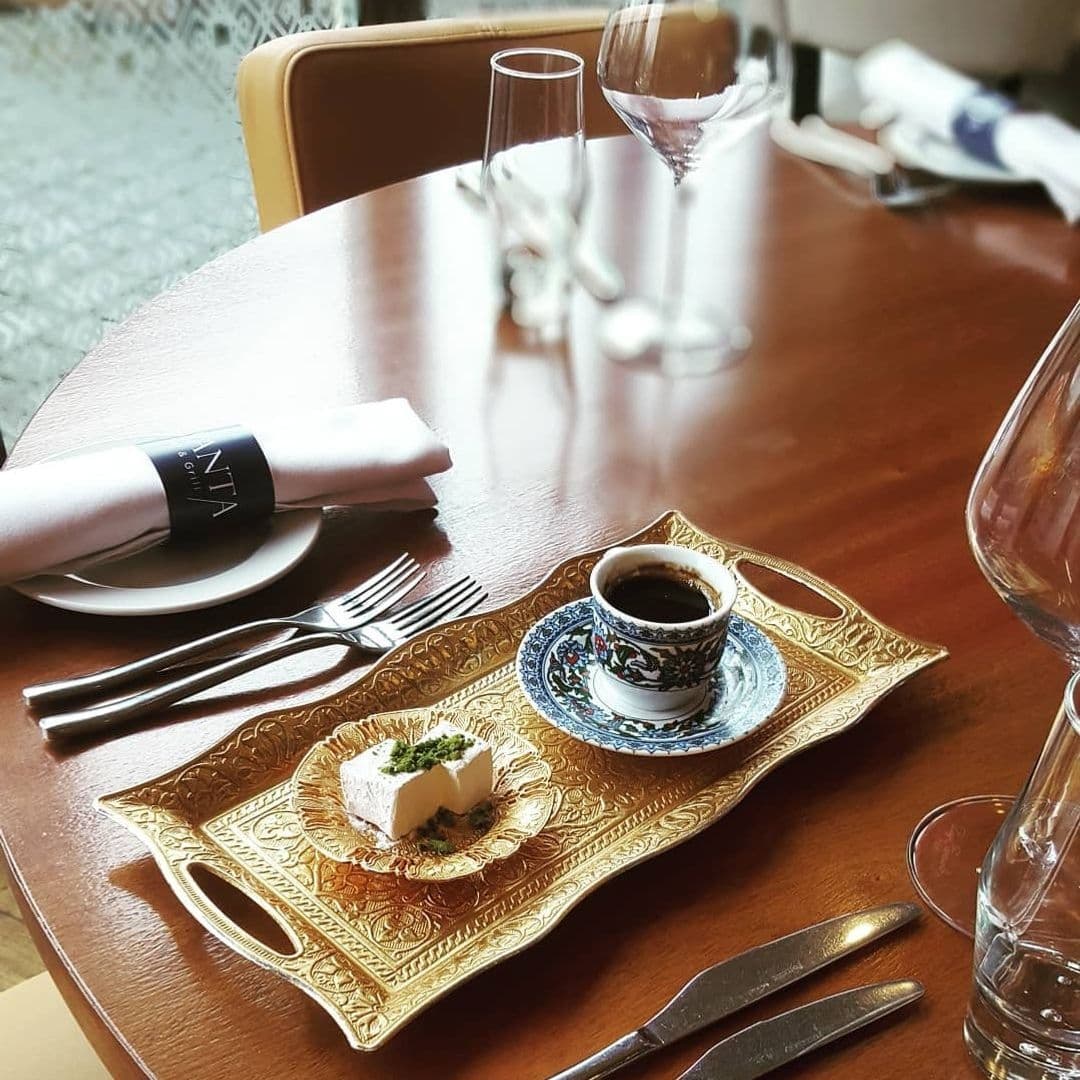 Lavanta Meze Bar & Grill Elegant Table Setting Turkish Coffee Dessert Elegant dining setup at Lavanta Meze Bar & Grill featuring a traditional Turkish coffee in a decorative cup on a golden tray, accompanied by a small dessert, with a set table in the background.
