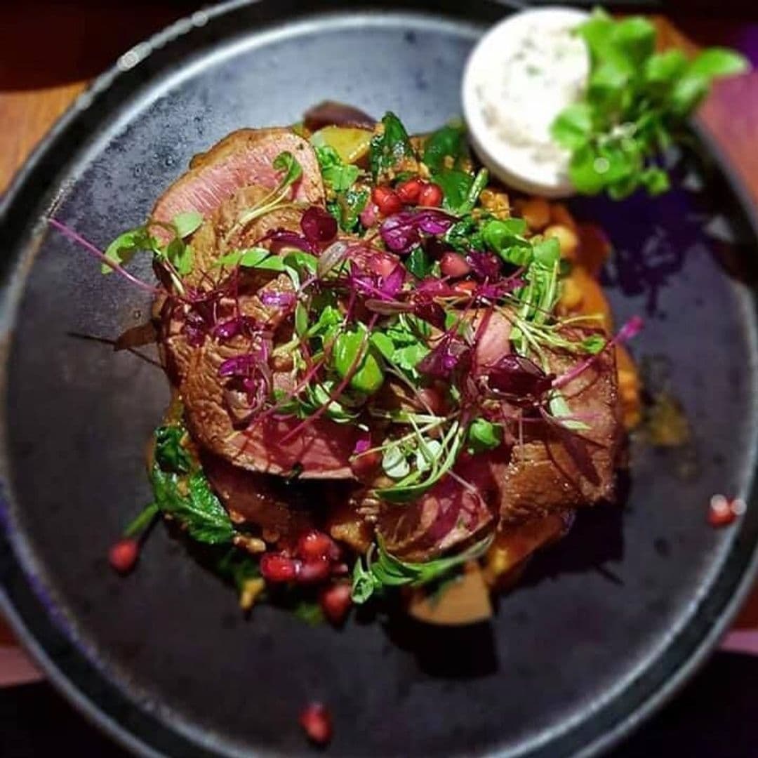 Lavanta Meze Bar & Grill Gourmet Roast Beef Pomegranate Salad Gourmet dish at Lavanta Meze Bar & Grill featuring thinly sliced roast beef topped with fresh herbs, pomegranate seeds, and microgreens, served with a side of creamy sauce on a dark plate.
