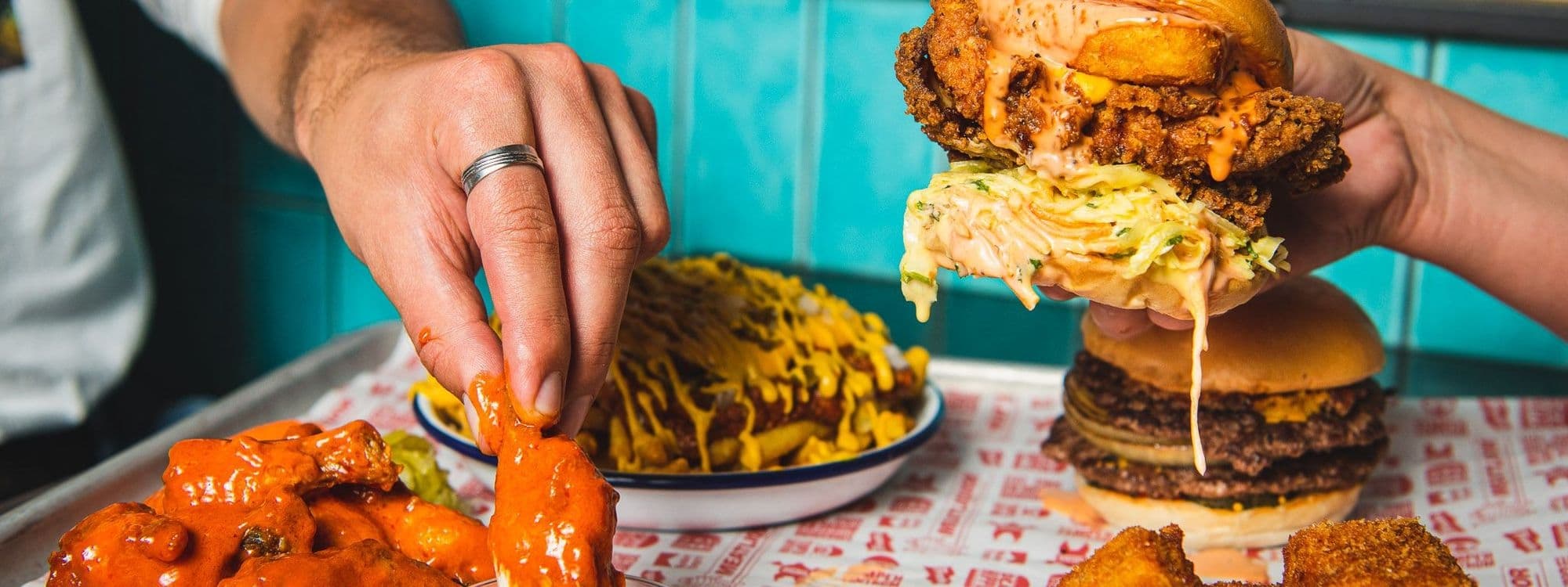 Alt text: "A tantalizing display of MEATliquor's indulgent cuisine, featuring a person's hands holding a towering burger dripping with sauce, alongside a plate of spicy buffalo wings and cheese-covered fries."
