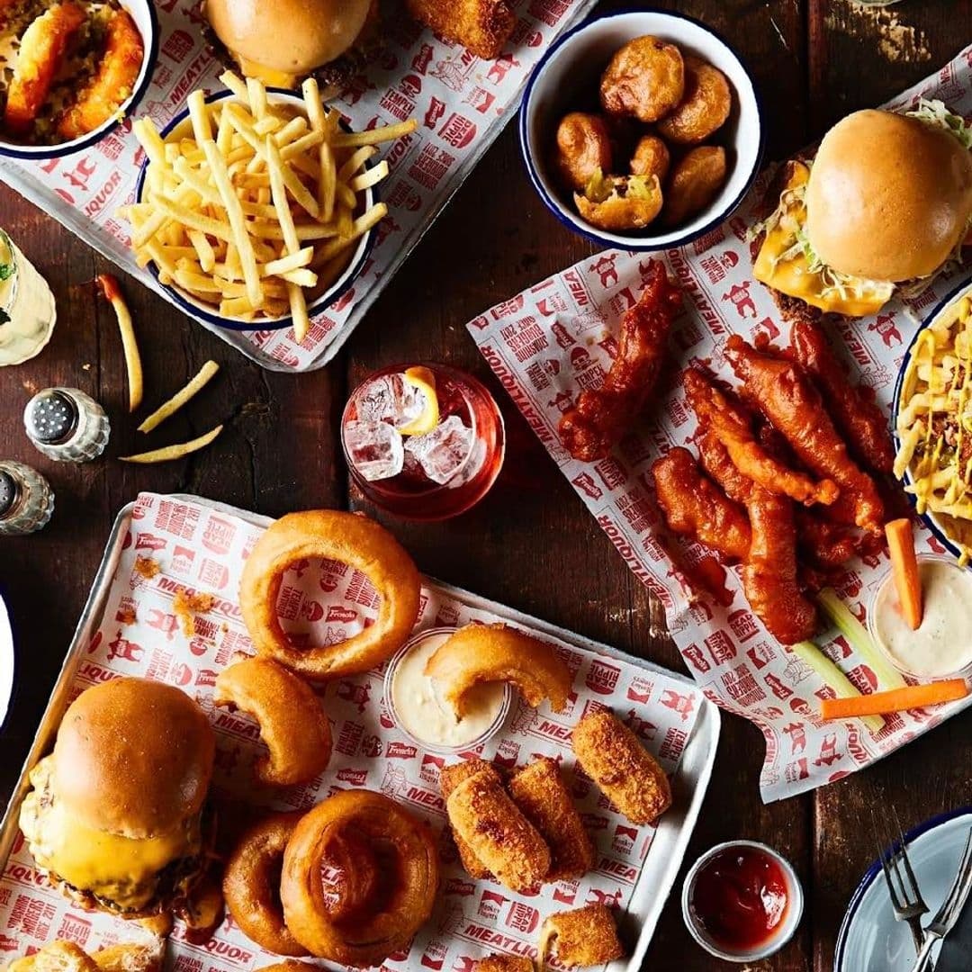 MEATliquor_Burgers_Fries_Cocktails_ComfortFood Assorted American-style fast food dishes on a table at MEATliquor, featuring burgers, fries, onion rings, chicken wings, and mozzarella sticks, accompanied by dipping sauces and a cocktail.