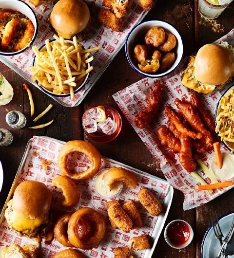 MEATliquor_Burgers_Fries_Cocktails_Feasting Assorted American-style fast food dishes on a table at MEATliquor, featuring burgers, fries, chicken wings, onion rings, and mozzarella sticks, with condiments and a cocktail.