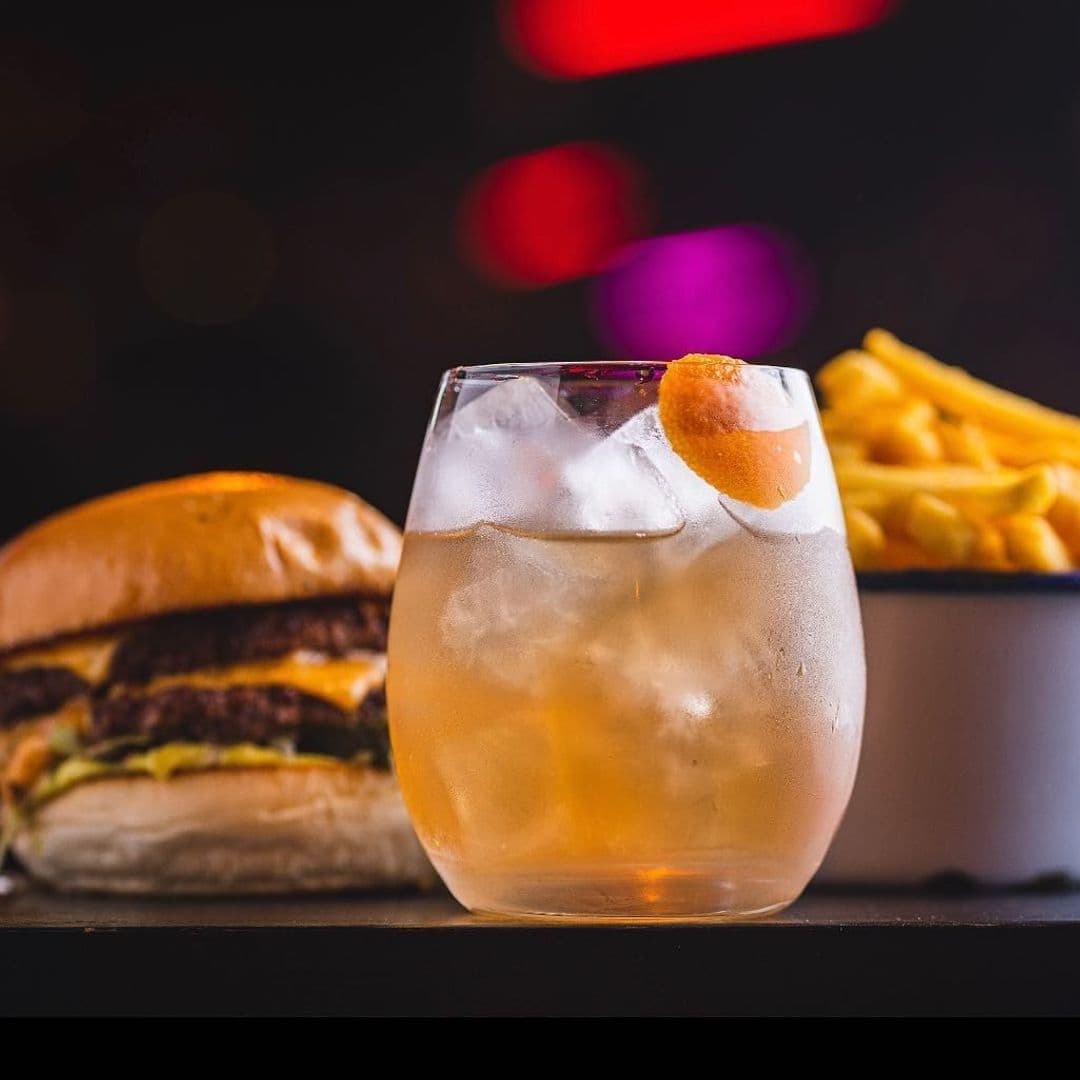 MEATliquor_Cocktail_Burger_Fries_Ambiance Cocktail with ice and garnish in the foreground, cheeseburger and fries in the background at MEATliquor.