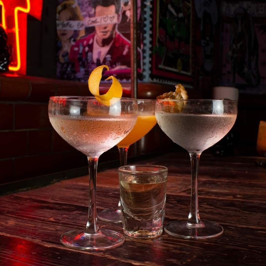 MEATliquor_Cocktails_NeonAmbience_BarScene Assorted cocktails on a bar table at MEATliquor with a neon-lit ambiance in the background.