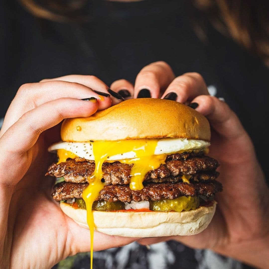 MEATliquor_Juicy_Cheeseburger_Melting_Cheddar Juicy double cheeseburger with melting cheese and pickles held in hands at MEATliquor restaurant.